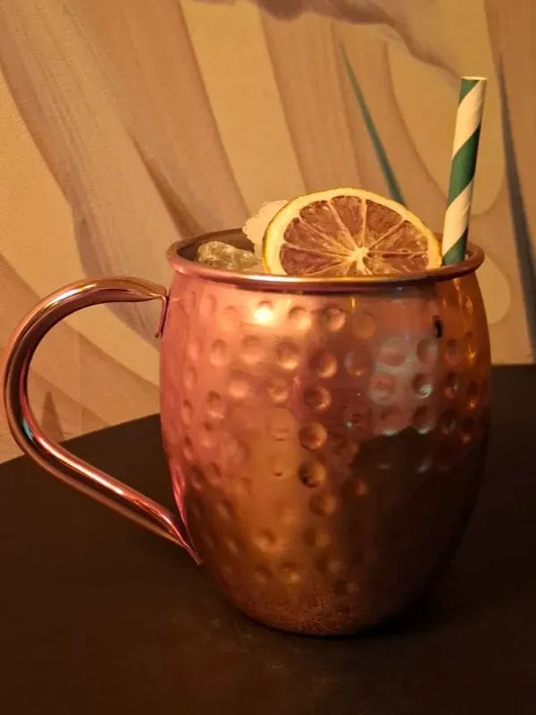 Un cóctel Moscow Mule servido en una taza de cobre martillado con una rodaja de cítricos secos y una pajita de papel a rayas 