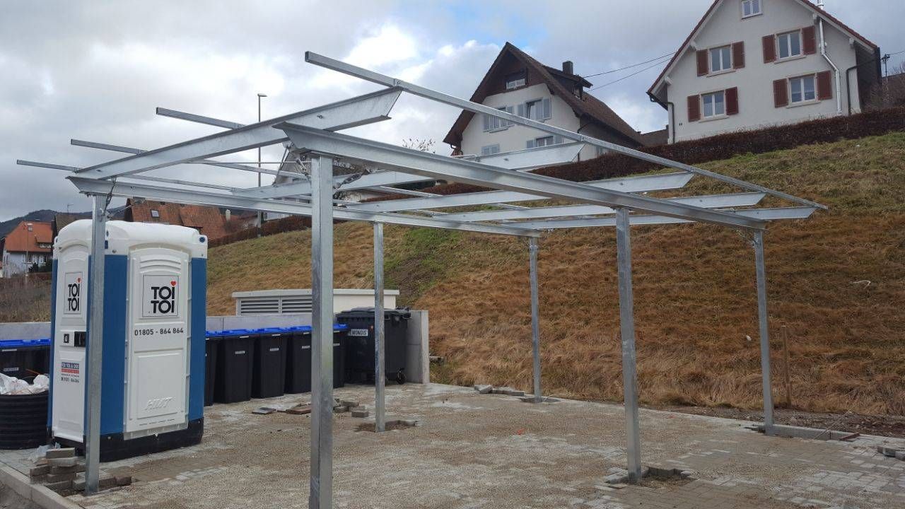 Eine tragbare Toilette steht unter einer Metallkonstruktion auf einem Parkplatz.