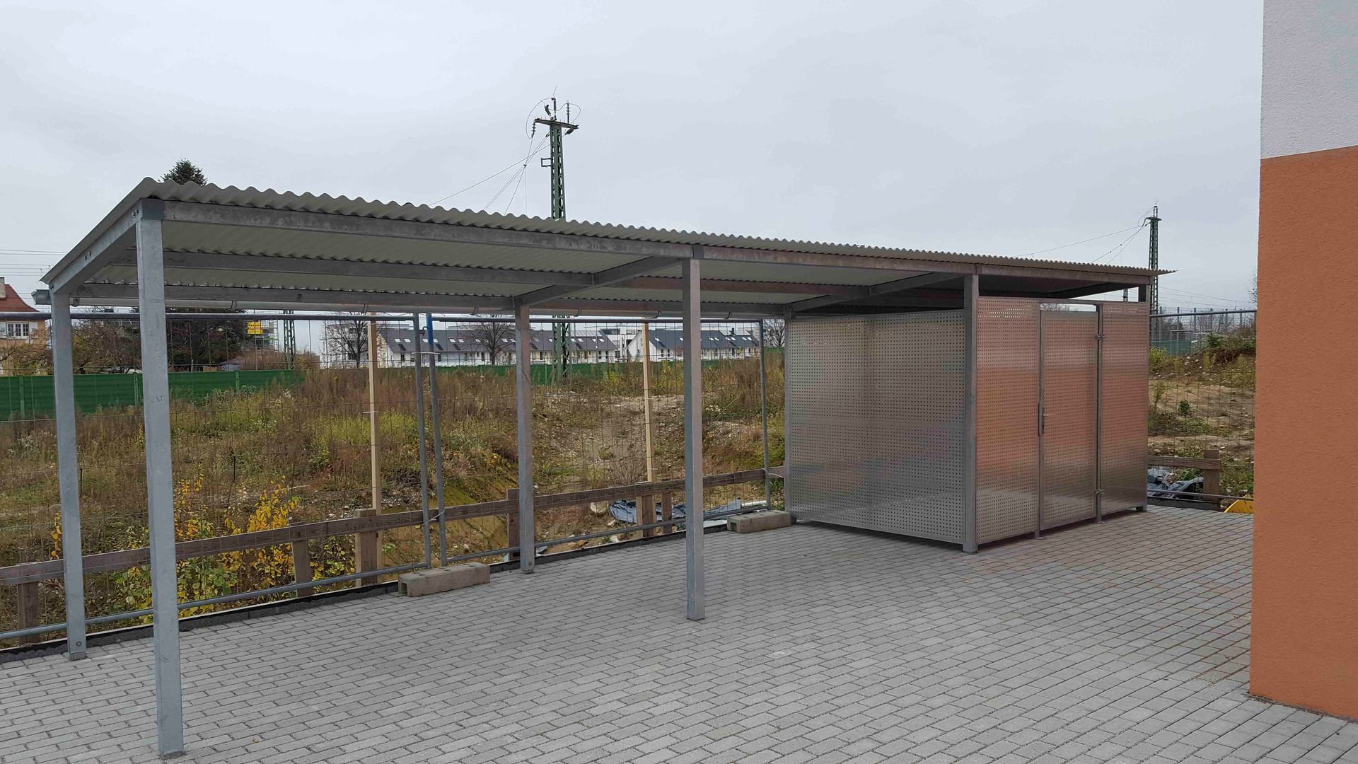 Ein Carport mit Metalldach steht auf einer gemauerten Auffahrt neben einem Gebäude.