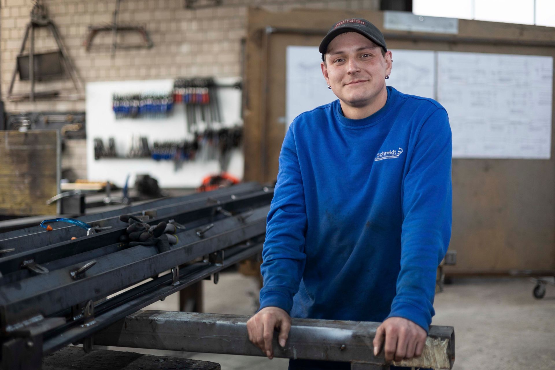 Ein Mann in einem blauen Pullover lehnt an einer Metallstange in einer Fabrik.