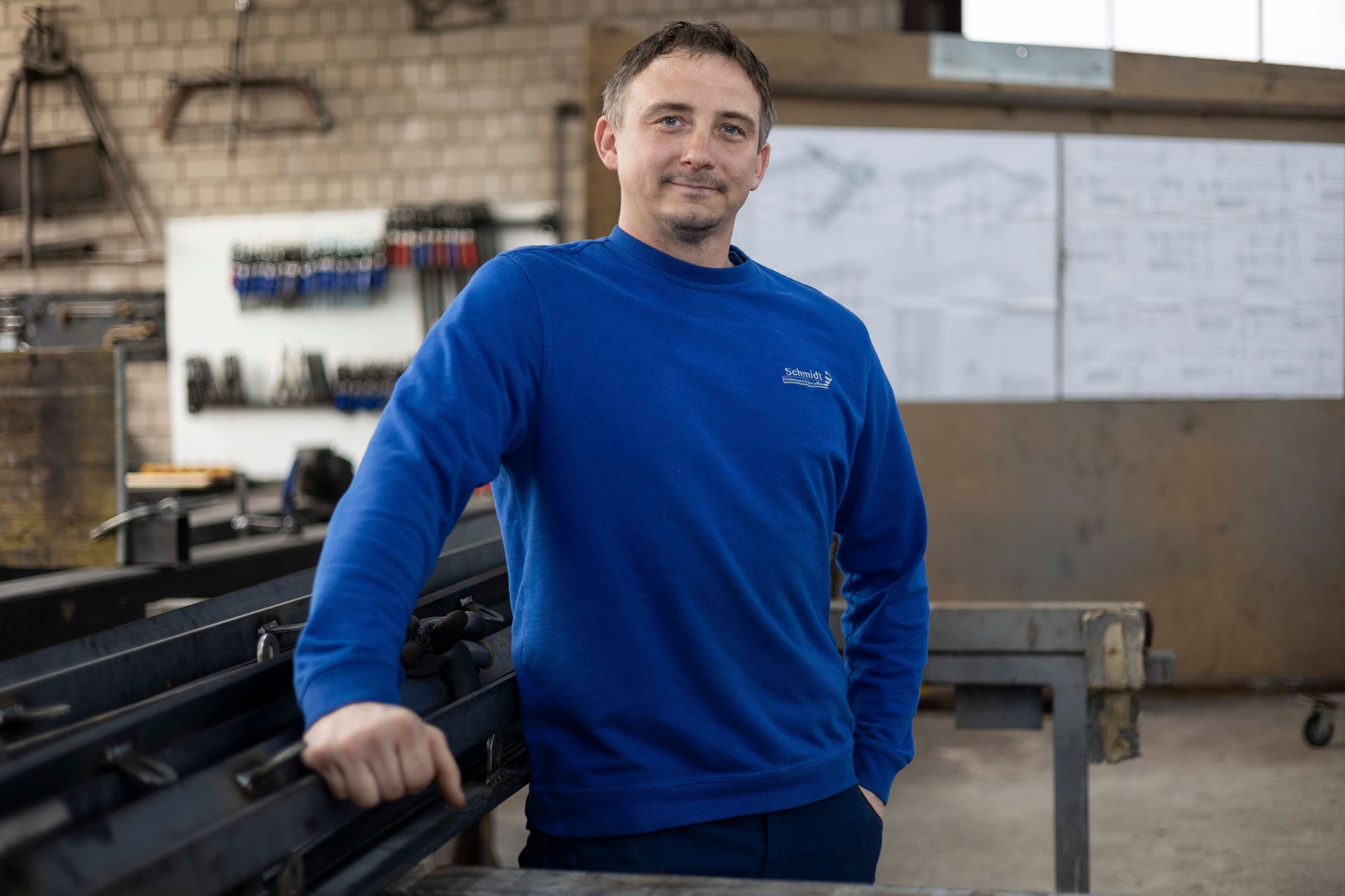 Ein Mann in einem blauen Pullover steht mit den Händen in den Taschen in einer Fabrik.