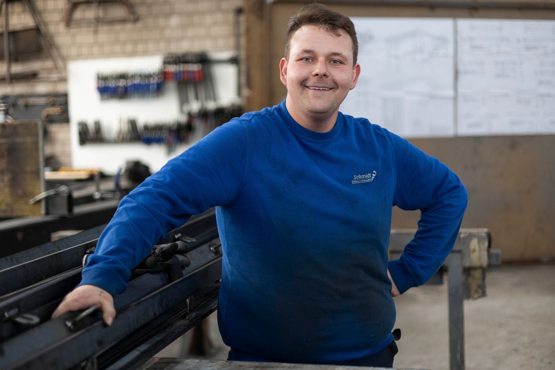 Ein Mann in einem blauen Pullover steht in einer Fabrik und hat die Hände in die Hüften gestemmt.