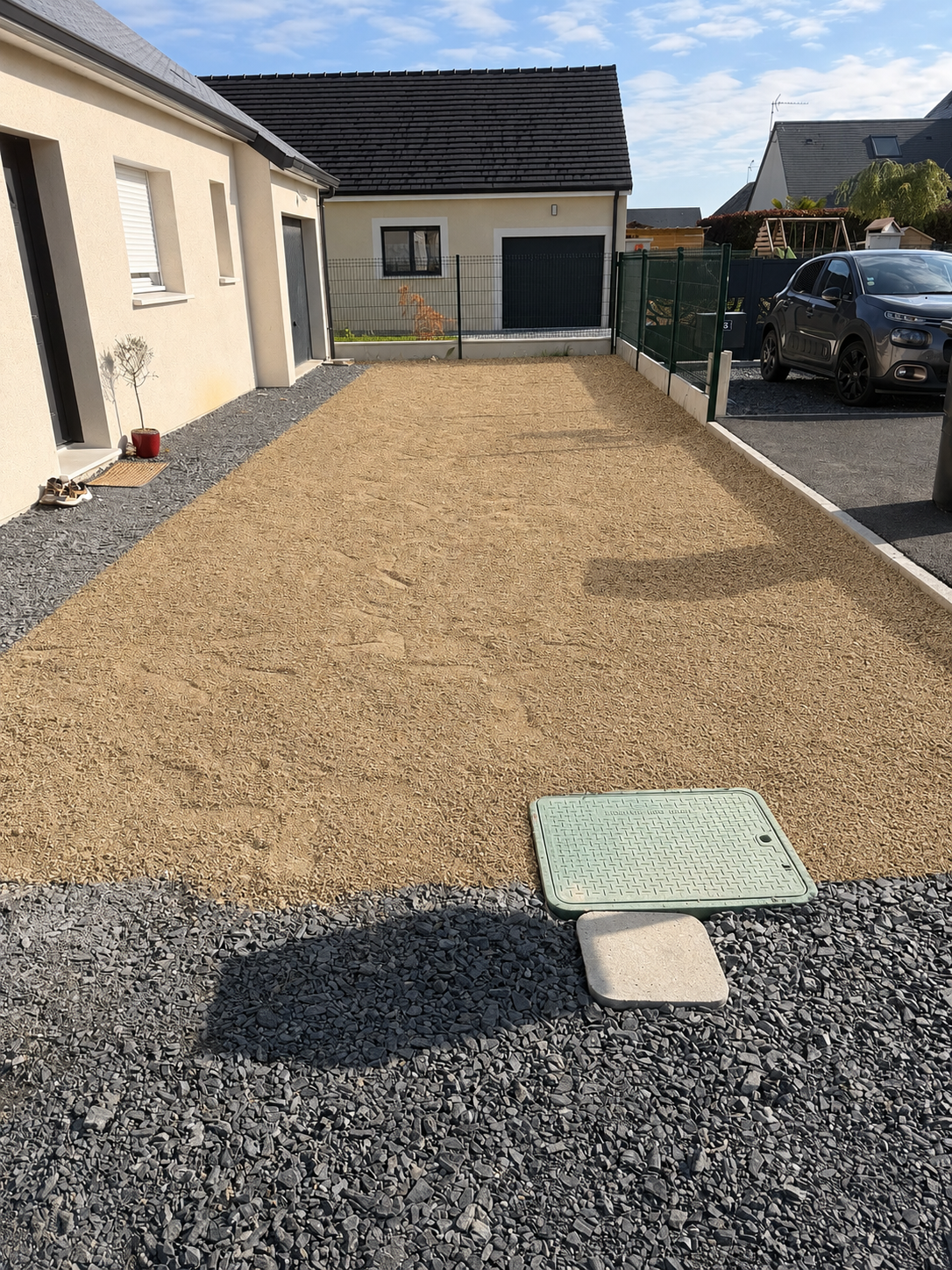 Allée de gravier ensoleillée à côté d'une maison et d'un garage, avec des voitures garées sur la droite.