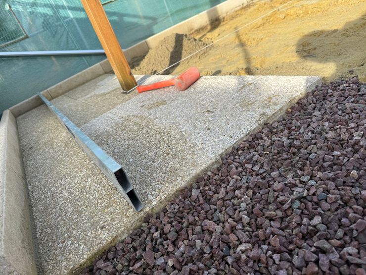 Finition d’aménagement extérieur avec dalle béton, gravier rouge et règle métallique de nivellement.
