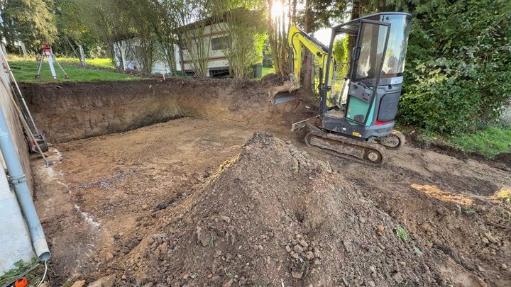 Une excavatrice nivelle la terre sur un chantier de terrassement, à côté d'une maison et d'arbres.