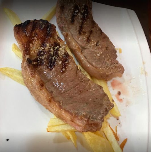 Dos filetes de ternera sellados a la parrilla, sazonados y servidos sobre una cama de patatas fritas finas y doradas en un plato.