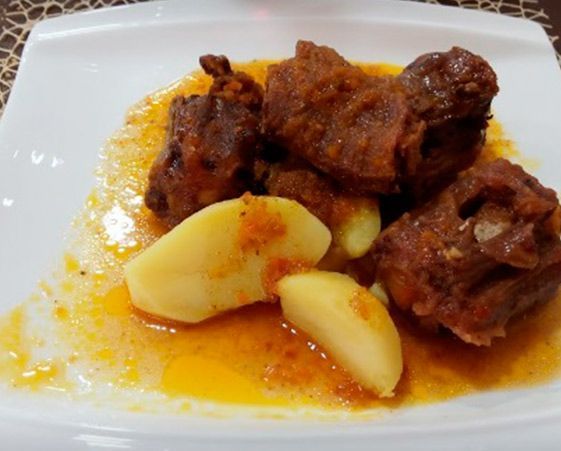 Un plato de trozos de carne estofada servidos en una salsa rica de color naranja con varios trozos de patata cocida.