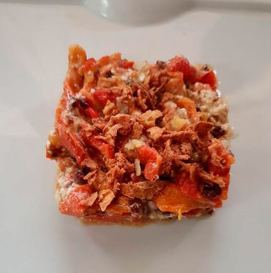 Una porción cuadrada de un guiso cremoso al horno, cubierto con tomates picados y cebollas fritas crujientes, servido en un plato blanco.