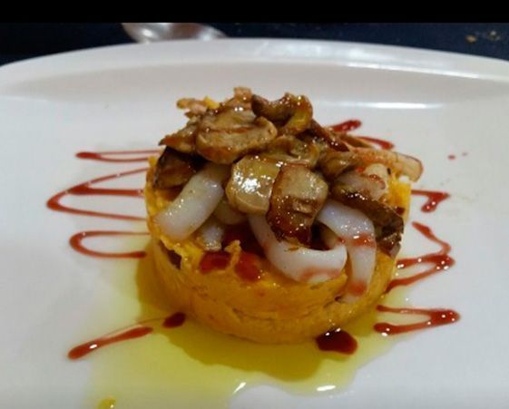 Un plato gourmet con una base moldeada de color naranja, cubierto con calamares blancos y champiñones salteados, y bañado con un glaseado rojo.