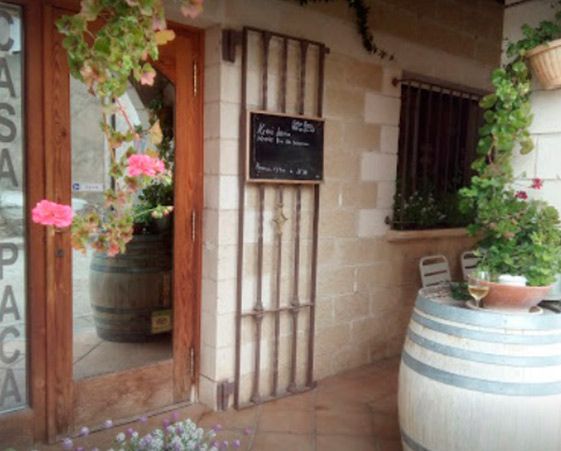 Entrada de un restaurante rústico con puerta de madera, pared de piedra, un pequeño cartel de pizarra y barriles de vino con plantas.