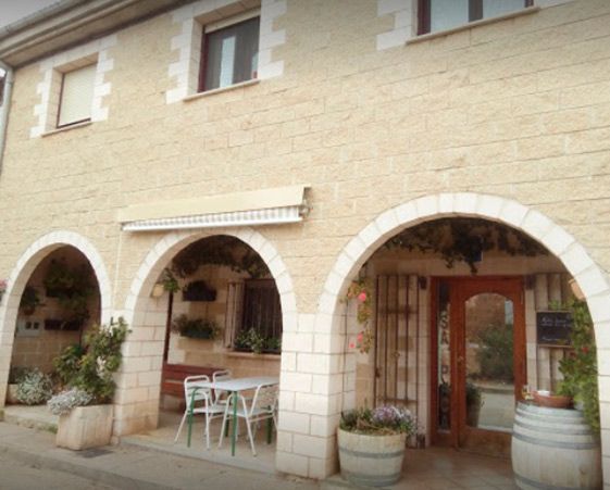 Un edificio de piedra con tres entradas arqueadas, asientos al aire libre y plantas en macetas bajo una fachada de color claro.