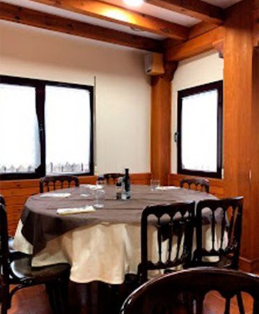 Una mesa redonda con un mantel beige y marrón está puesta para una comida en una habitación con vigas de madera oscura y ventanas enmarcadas.