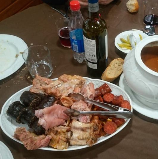 La mesa está puesta con una gran fuente de embutidos variados, entre los que se incluyen morcilla y chorizo, junto a una olla para sopa.
