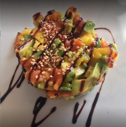 Una pila redonda de aguacate, mango y salmón cortados en dados, cubierta con semillas de sésamo y rociada con un glaseado balsámico oscuro.