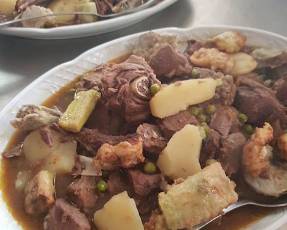 Primer plano de un guiso abundante con trozos de carne, patatas y guisantes en un caldo marrón, servido en una fuente ovalada blanca.