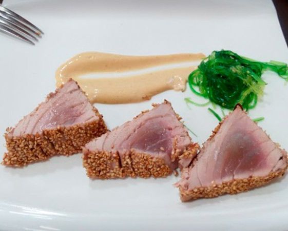 Tres filetes de atún con costra de sésamo dispuestos en un plato con un poco de salsa y una porción de ensalada de algas verdes.