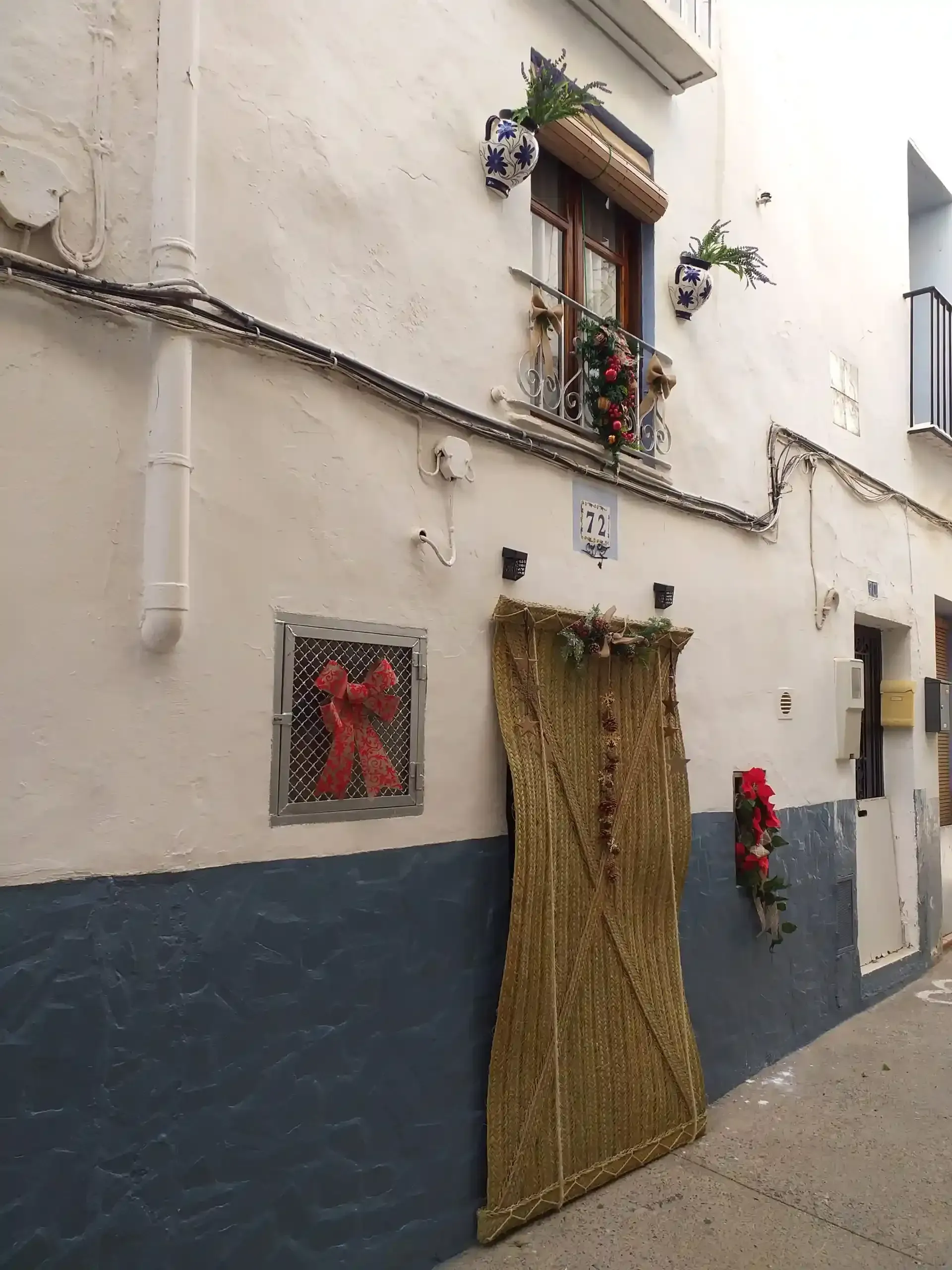 Calle estrecha con una pared blanca y azul, una ventana pequeña y una puerta decorada con un tapiz tejido y adornado con flores.