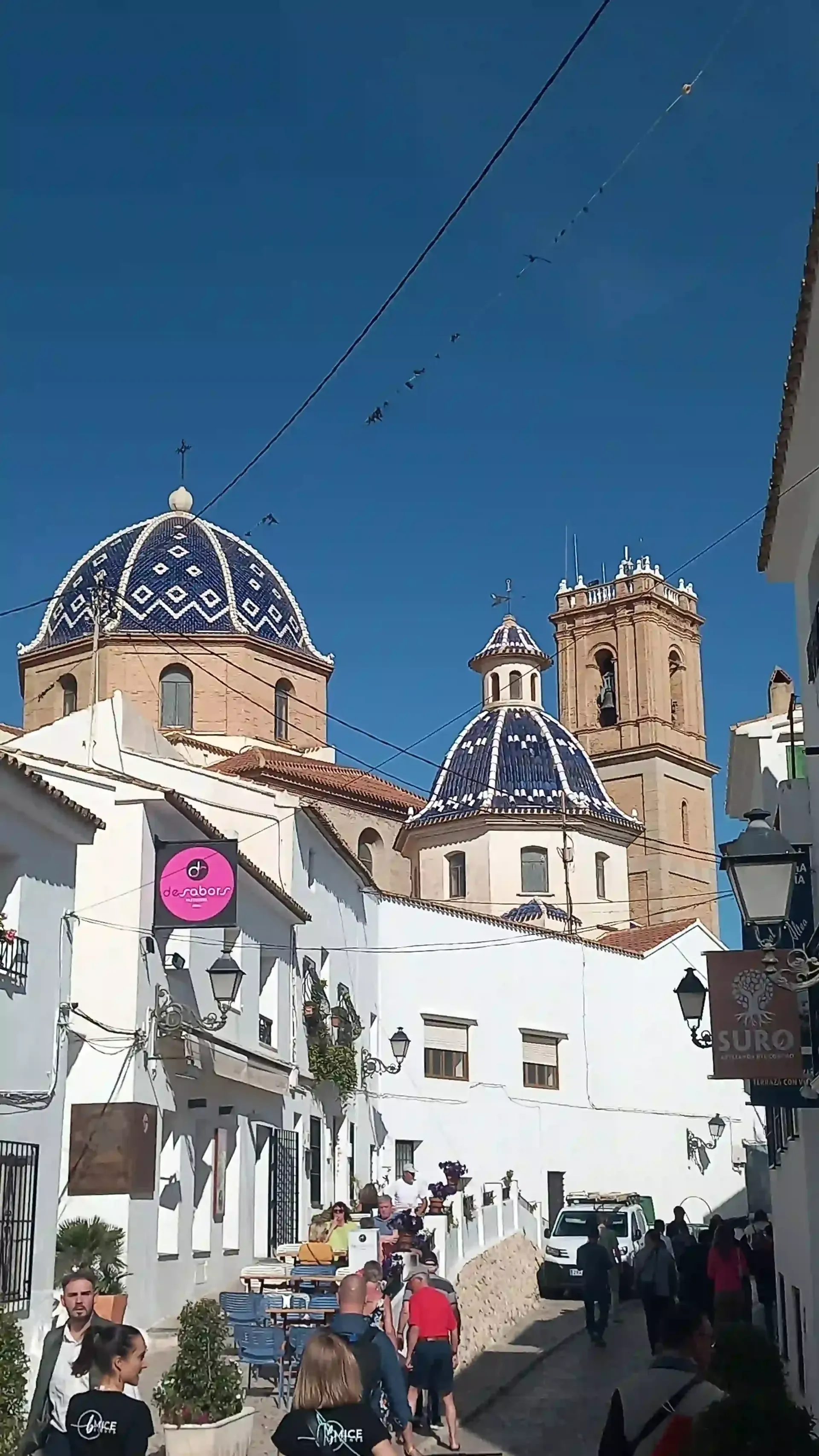Calle blanca bañada por el sol, con torres de iglesias de cúpulas azules y gente paseando por una estrecha callejuela del casco antiguo.