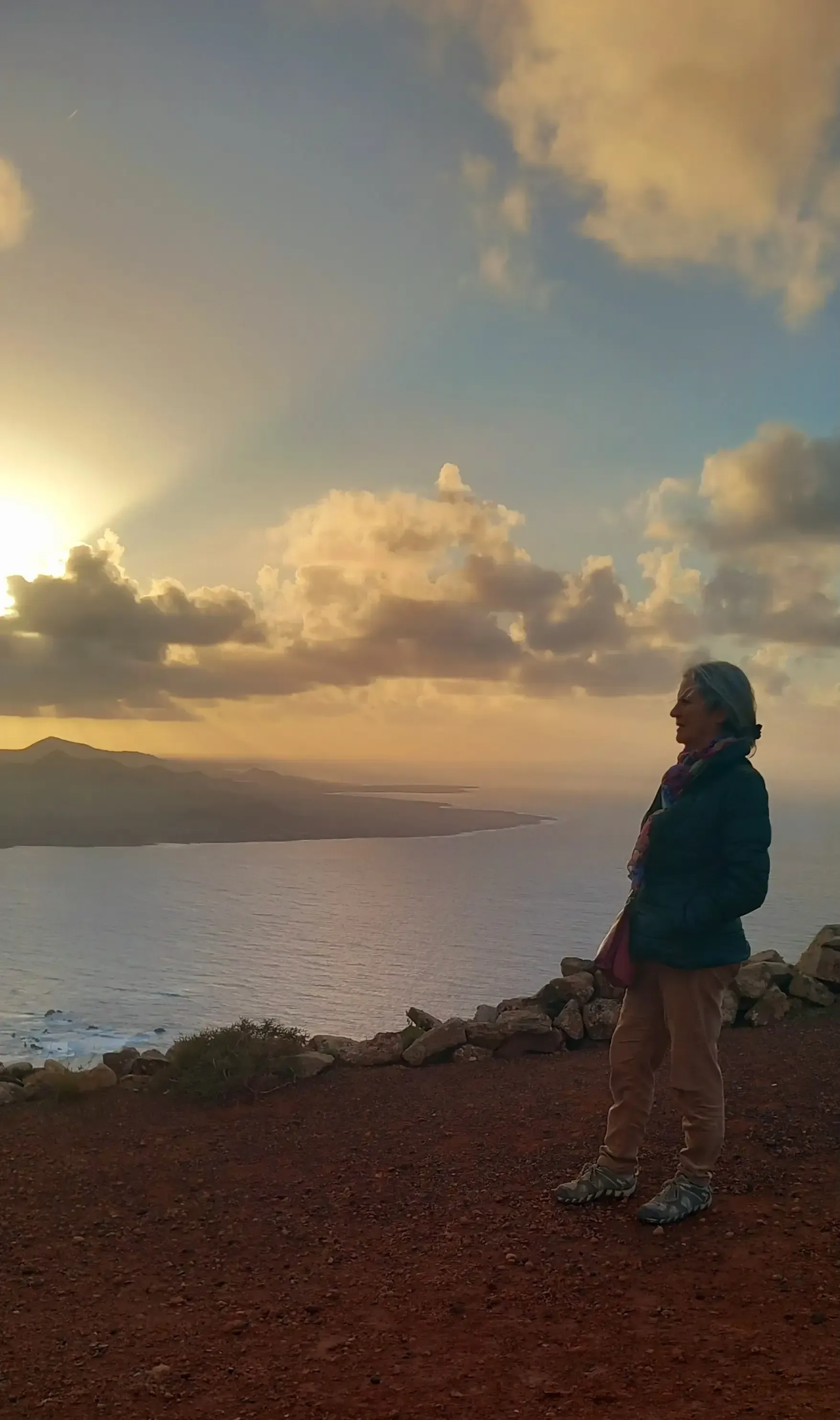 Una persona de pie en una orilla rocosa al atardecer, contemplando las aguas tranquilas y el cielo nublado.