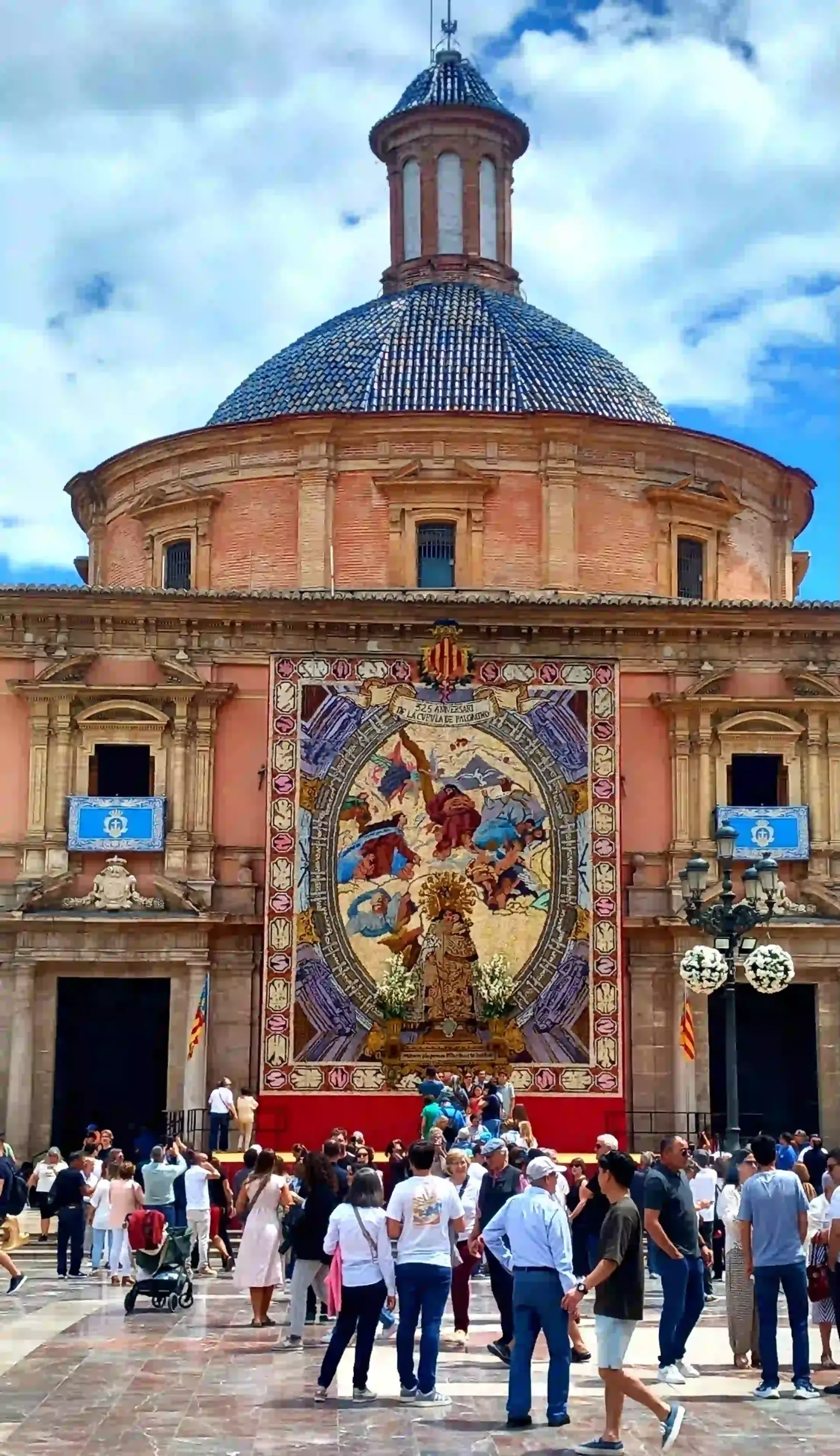 Multitud frente a un edificio histórico con un gran mosaico colorido y un tejado de cúpula azul.