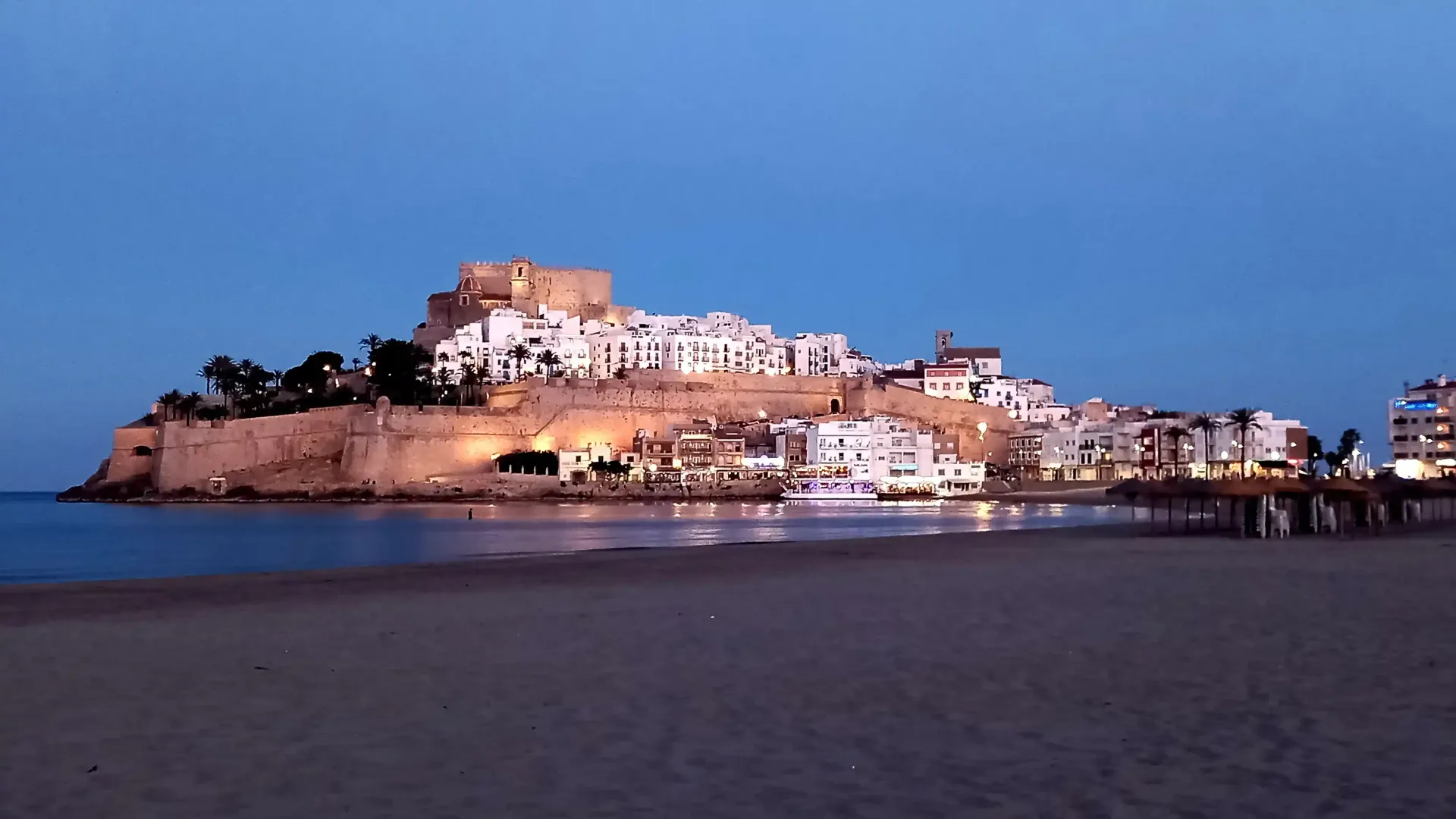 Pueblo costero iluminado sobre una colina rocosa al atardecer, reflejado sobre aguas tranquilas y oscuras.