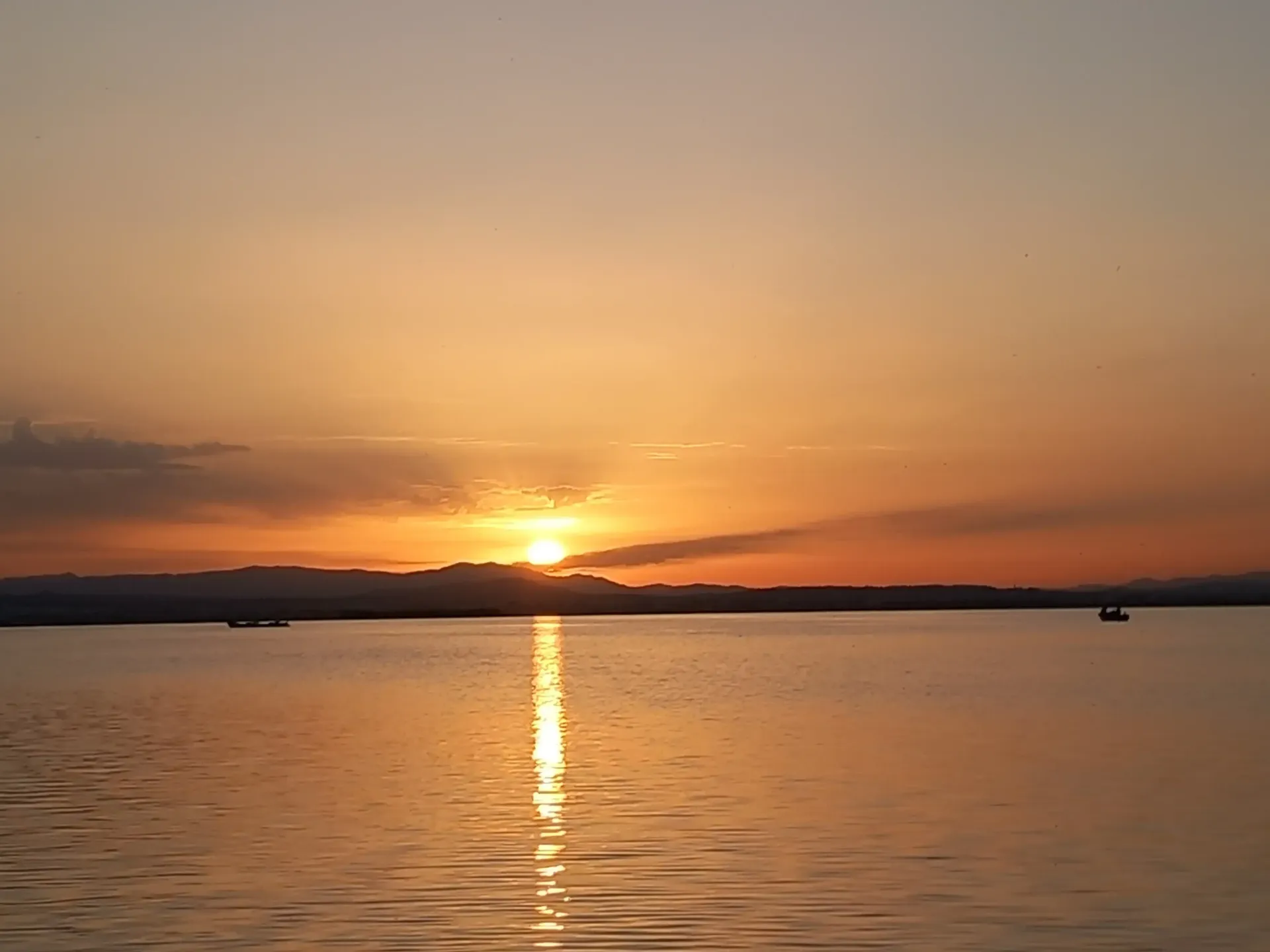 Puesta de sol sobre aguas tranquilas con un brillante reflejo y siluetas de la costa a lo lejos.
