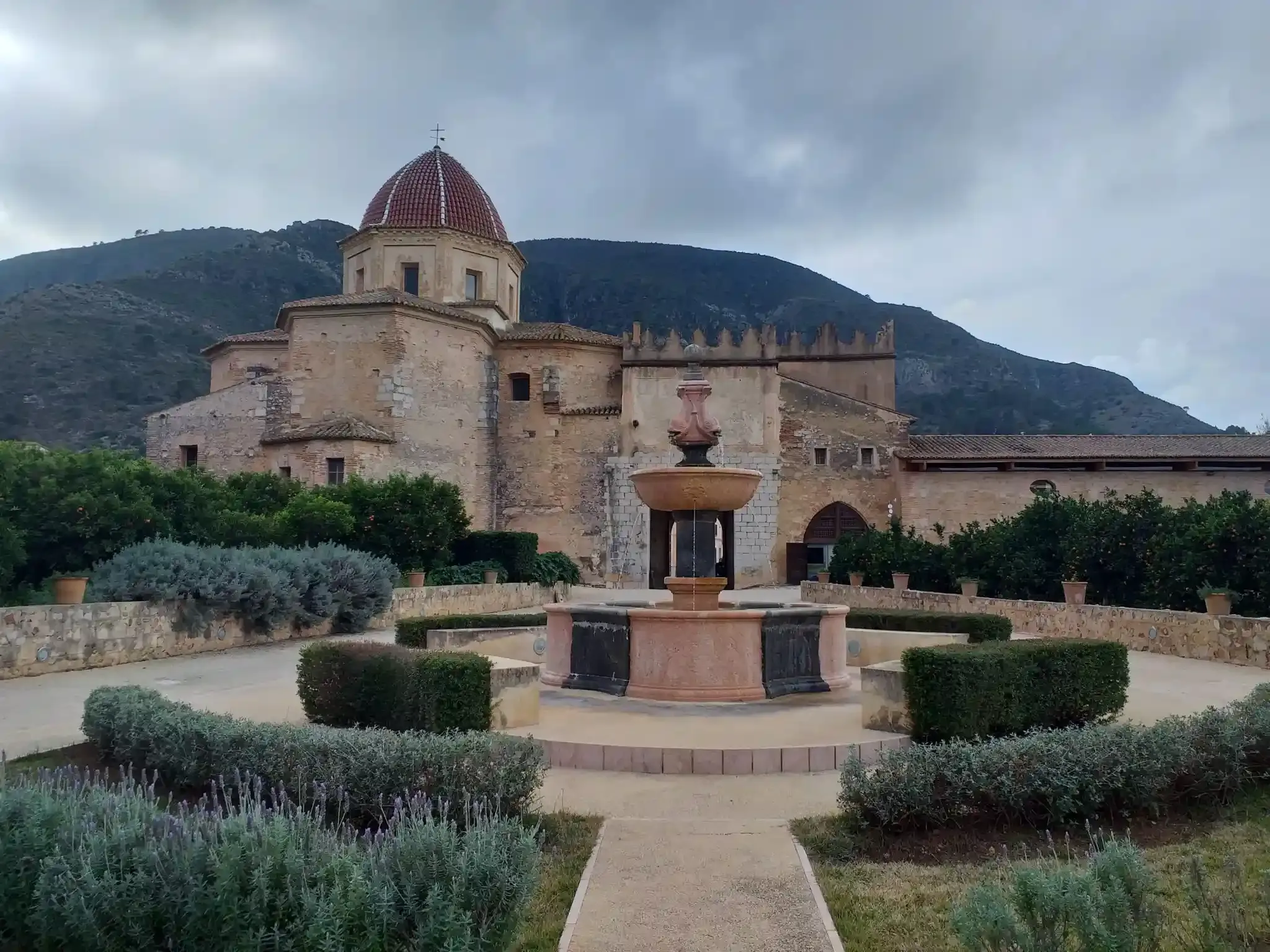 Iglesia histórica de piedra con cúpula y fuente en el patio, enmarcada por setos y montañas al fondo.