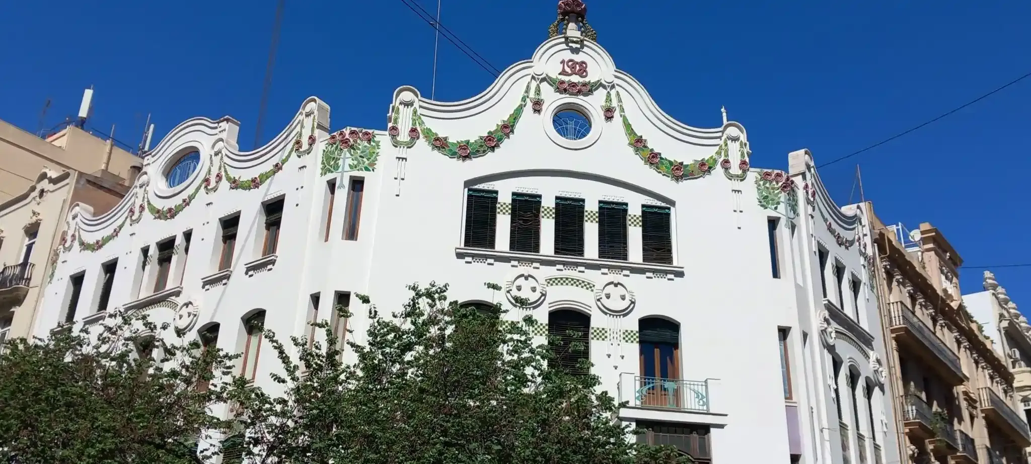 Edificio de apartamentos blanco y ornamentado con coloridos detalles en el tejado bajo un cielo azul claro.