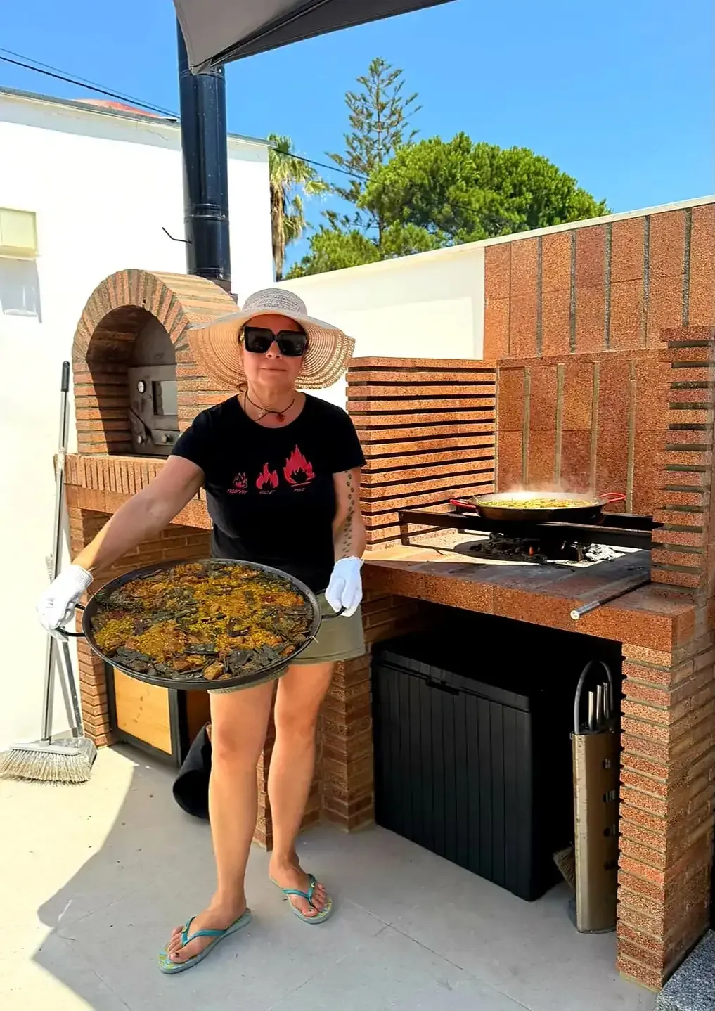 Una persona sostiene una gran bandeja de comida junto a un horno de ladrillos al aire libre en un patio soleado.