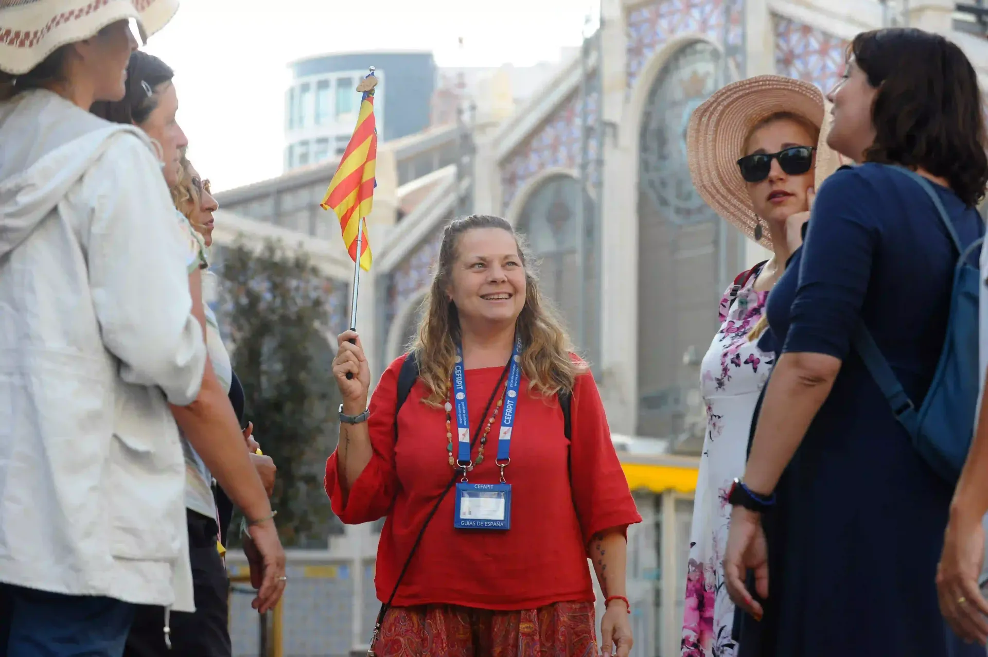 Un grupo de personas charlando al aire libre; una mujer con una camisa roja sonríe y sostiene una pequeña bandera.