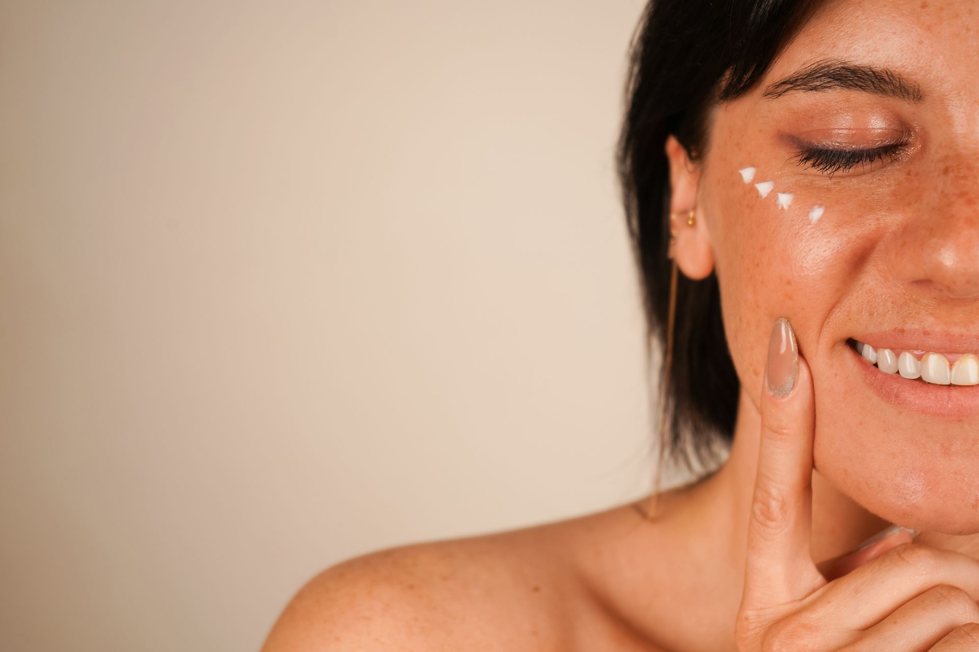 Femme appliquant une crème blanche sur sa joue, souriant sur un fond beige