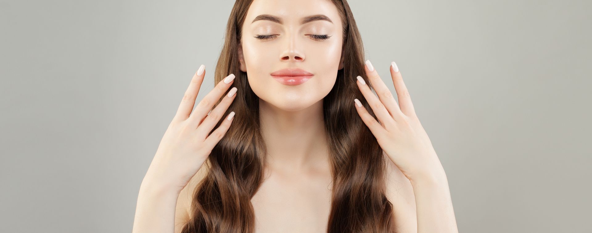 Femme aux longs cheveux bruns, les yeux fermés, se touchant le visage sur fond gris uni