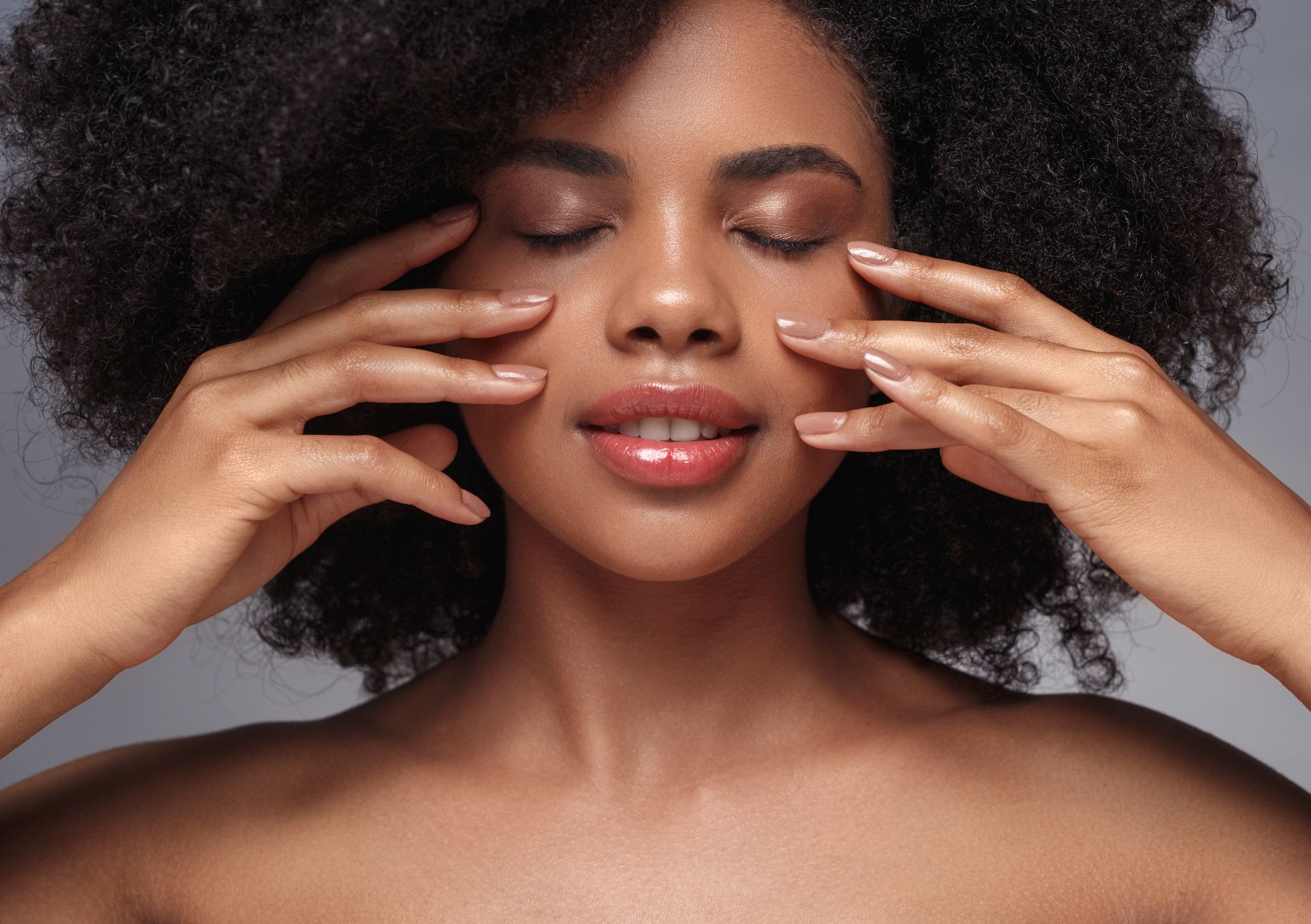 Femme aux cheveux bouclés appliquant une crème de soin sur ses joues, les yeux fermés sur fond gris