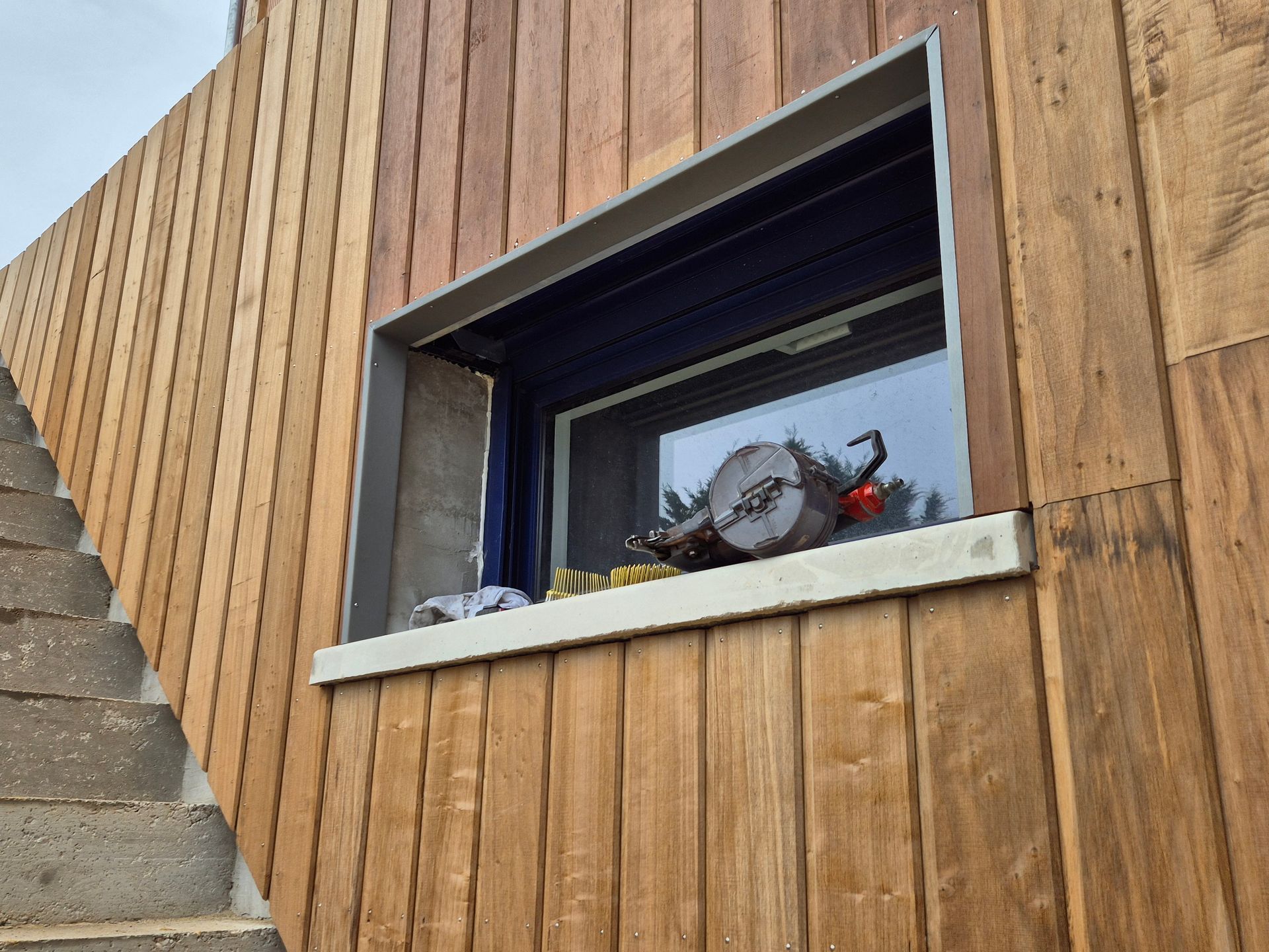 Bâtiment en bois avec fenêtre, tronçonneuse sur le rebord de la fenêtre. Escaliers en briques à gauche.