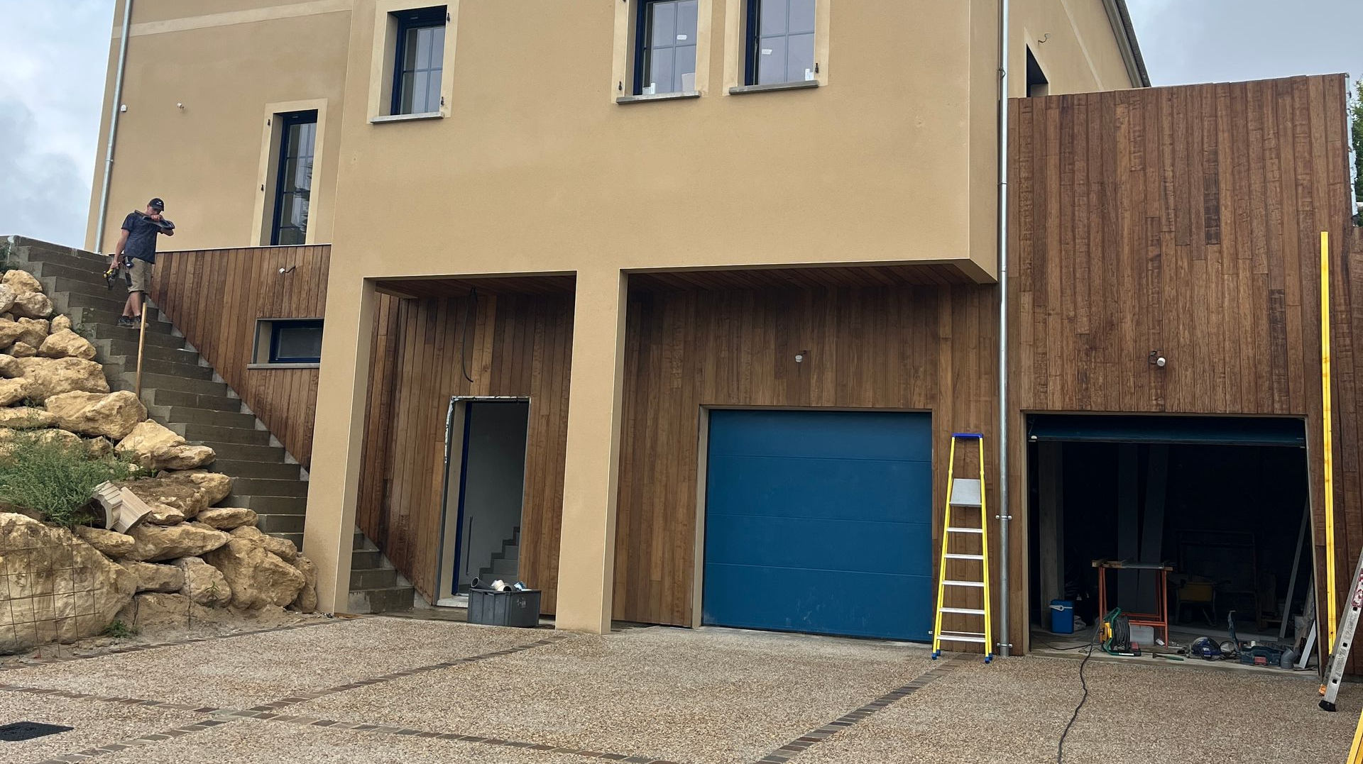 Maison à deux étages avec façade en stuc beige et éléments en bois. Porte de garage bleue, homme dans l'escalier, engins de chantier présents.