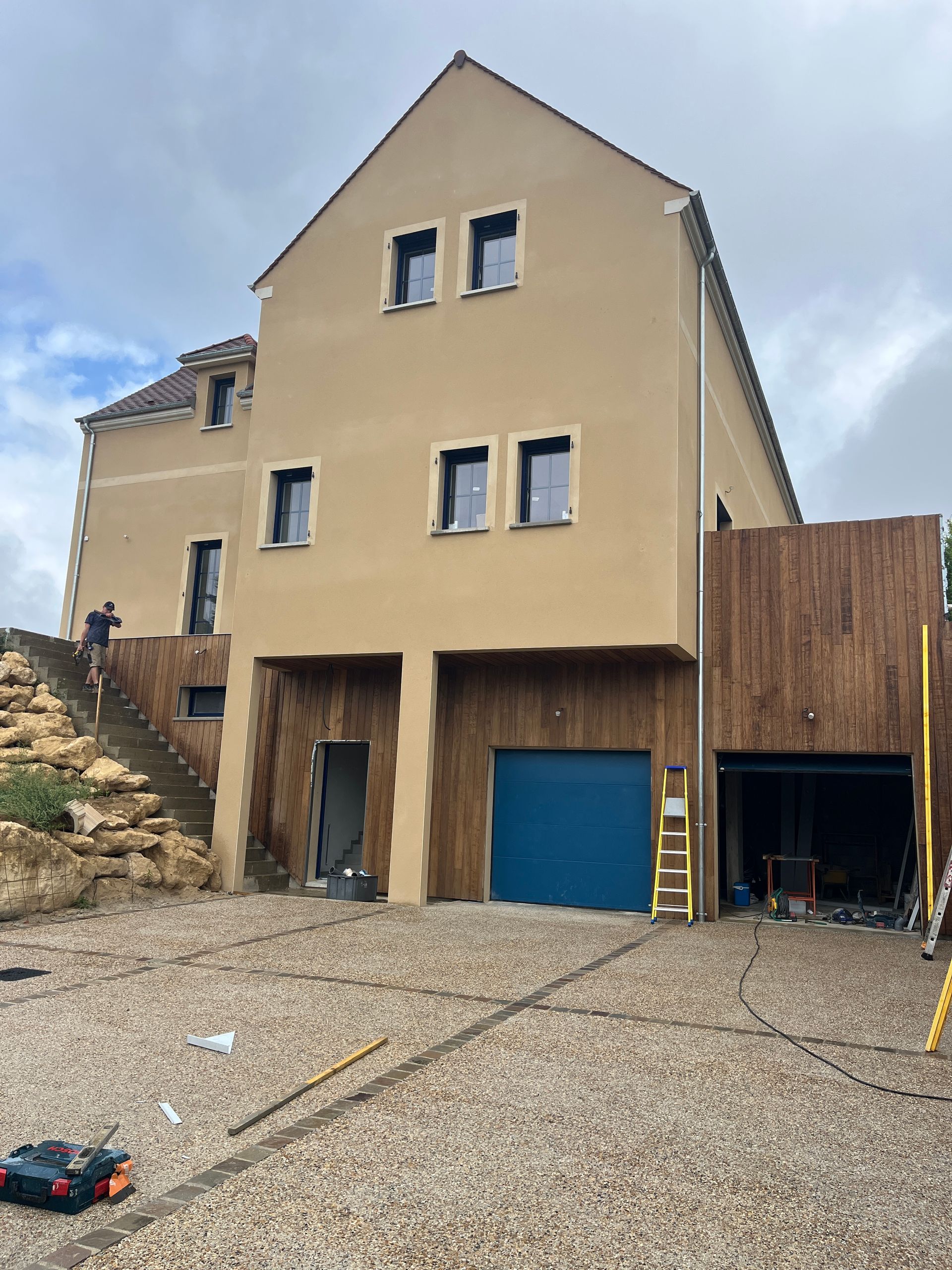 Maison à plusieurs étages de couleur beige avec porte de garage bleue et éléments en bois, allée en gravier.