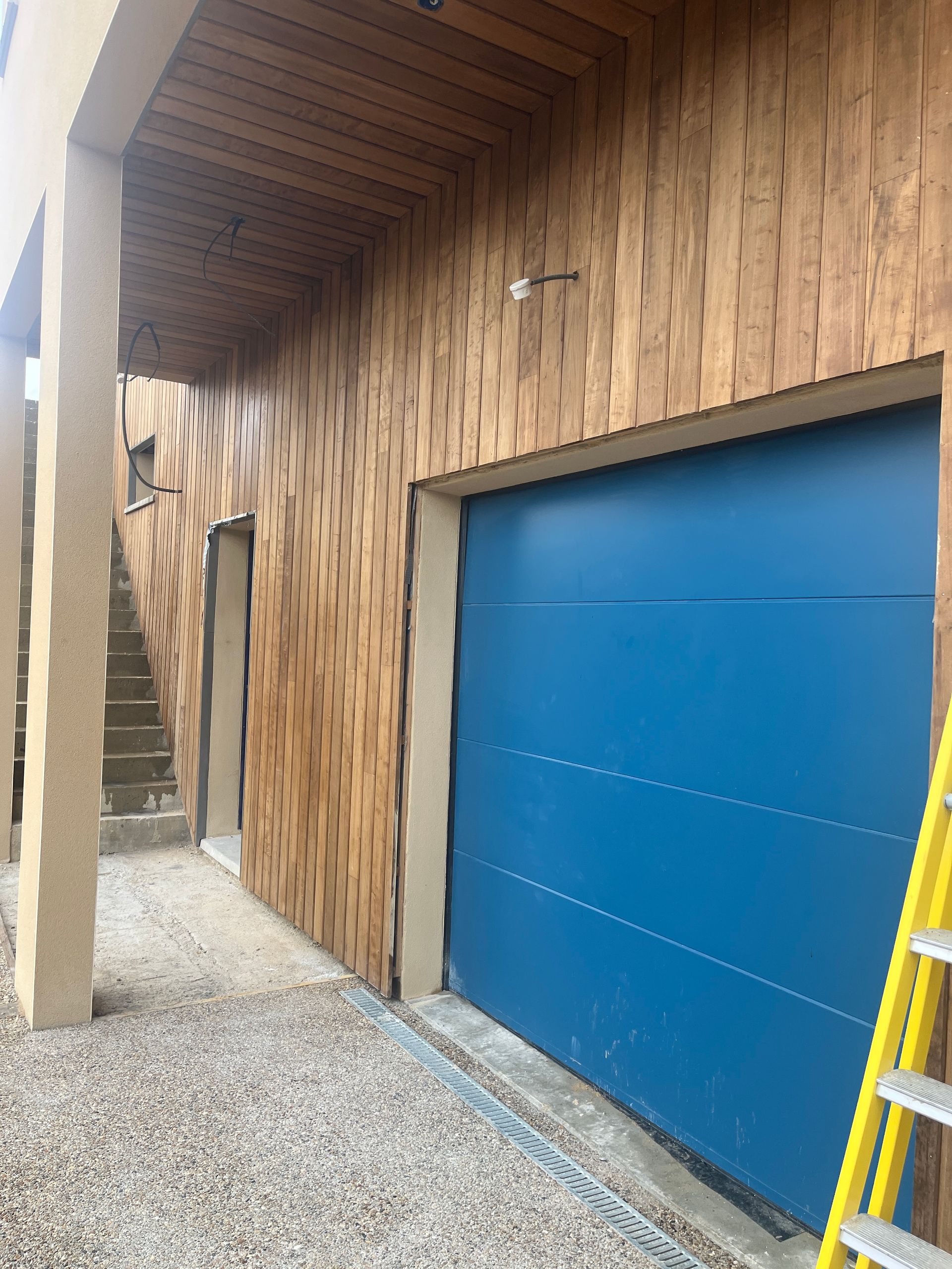 Porte de garage bleue sur un bâtiment à bardage en bois et escaliers en pierre, une échelle jaune est appuyée contre la droite.