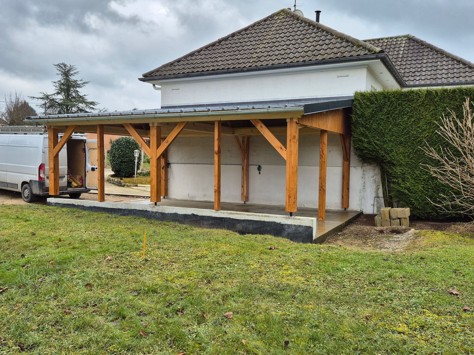 Un abri de voiture en bois est accolé à un bâtiment blanc ; une camionnette blanche, porte ouverte, est garée à proximité.
