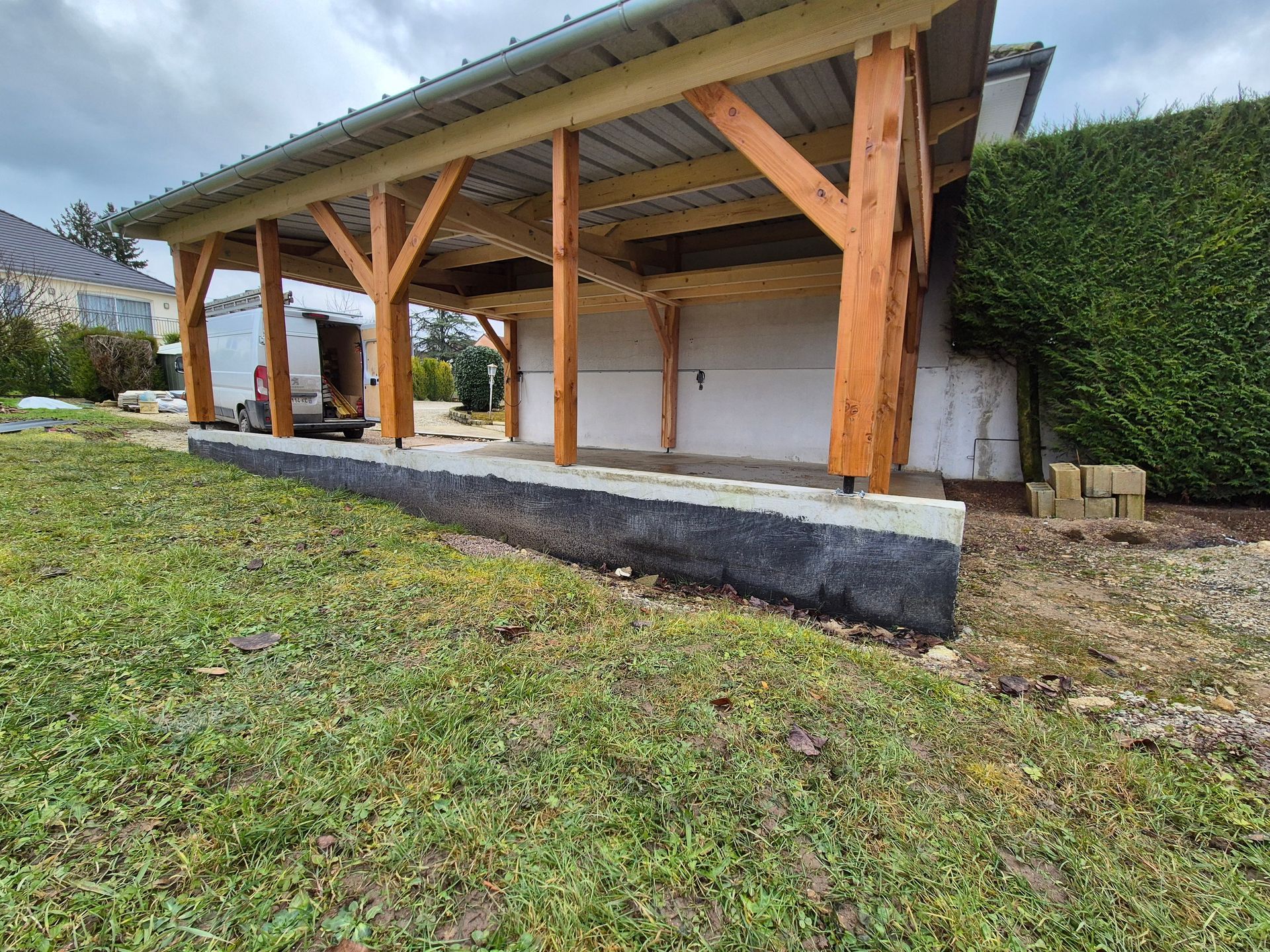 Abri de voiture en bois avec toit en métal, construit sur une base en béton.