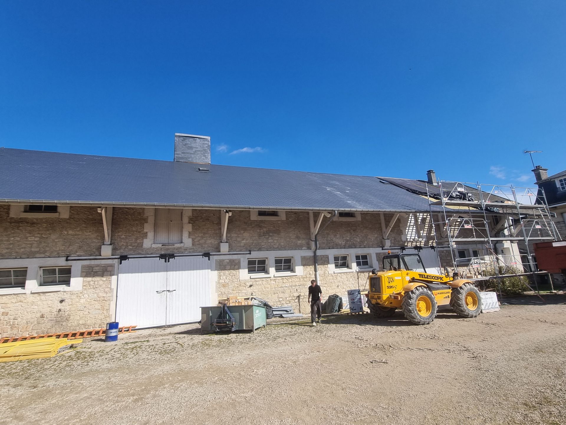 Bâtiment en construction, aux murs de pierre et au toit sombre ; un véhicule de chantier jaune est présent.