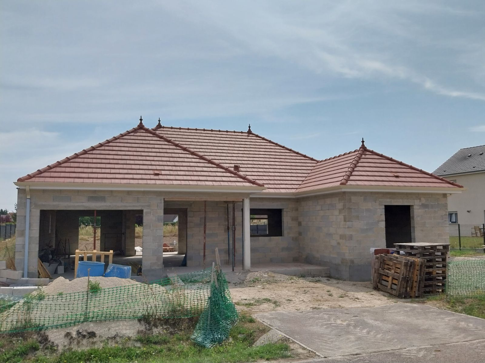 Maison en construction avec un toit de tuiles rouges et des murs en parpaings, par une journée ensoleillée.
