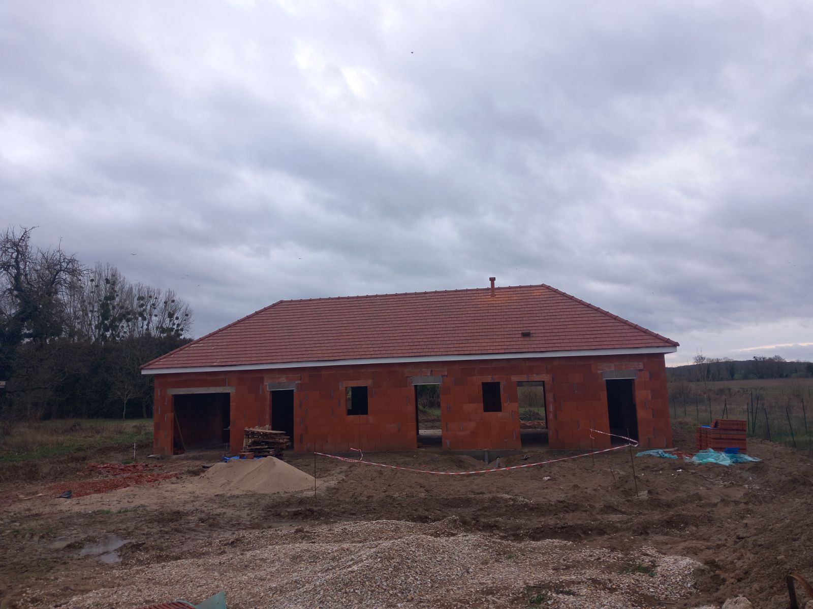 Maison en briques inachevée, au toit de tuiles rouges, sous un ciel couvert. Matériaux de construction et terre au premier plan.