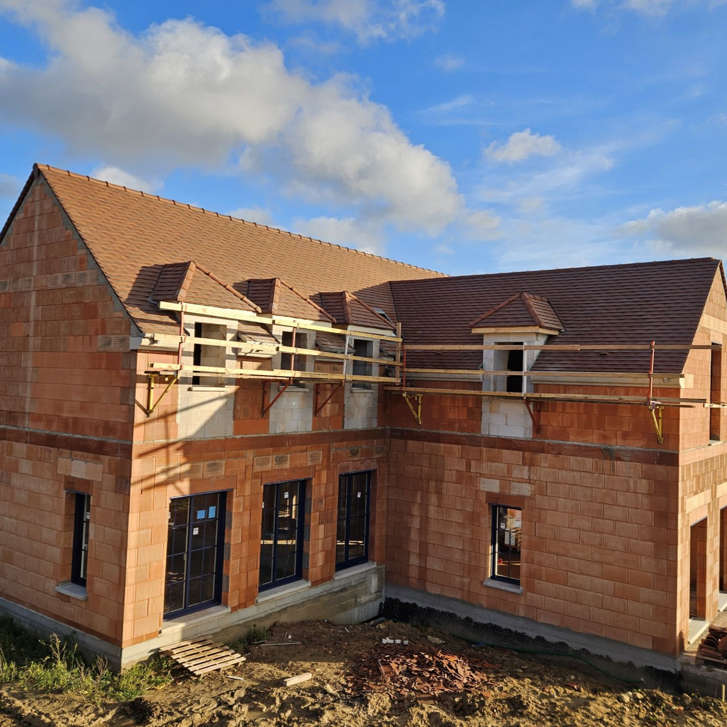 Construction d'une maison en briques de deux étages avec un toit de tuiles brunes et des échafaudages temporaires.