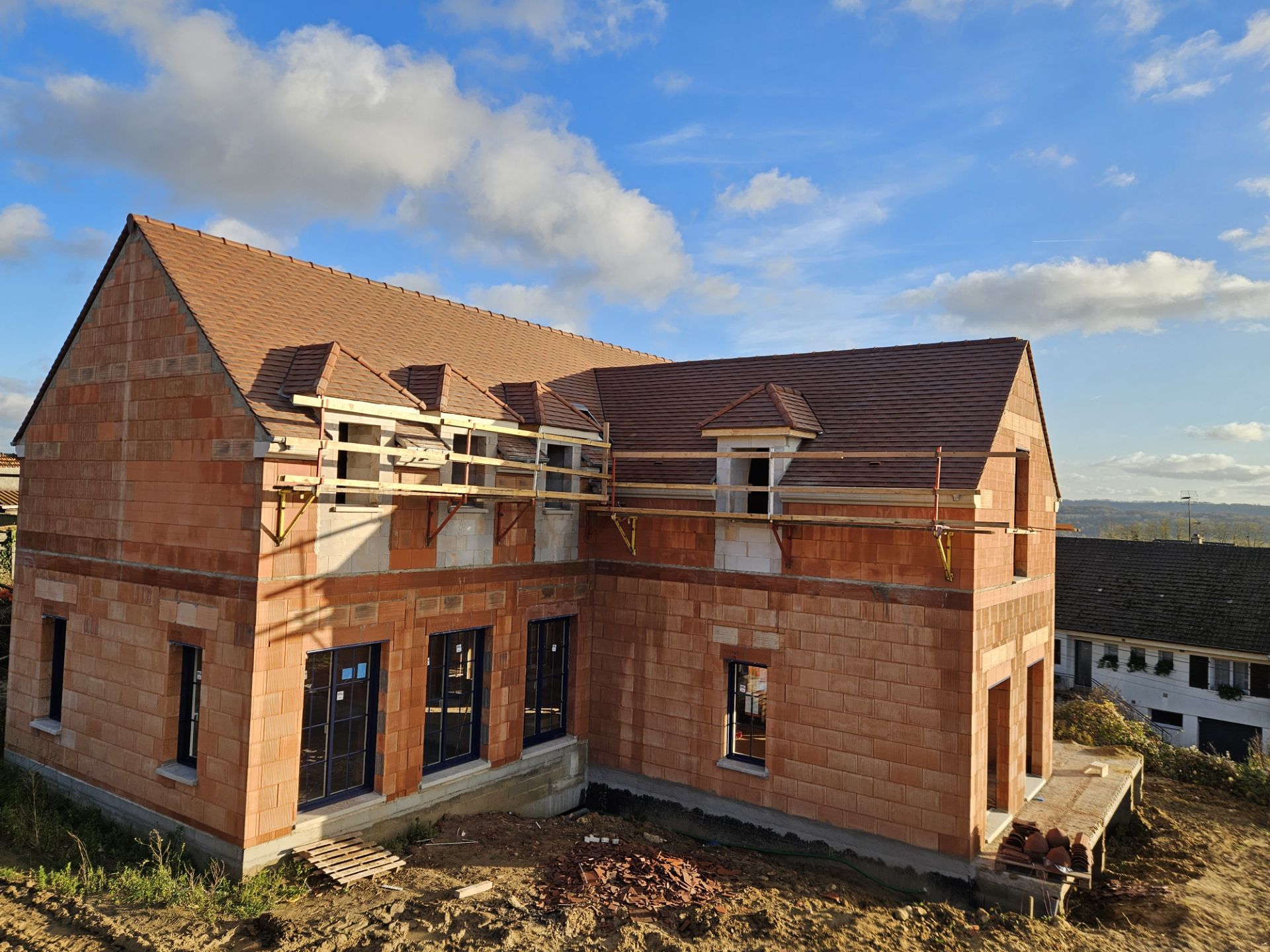 Maison en construction, façade en briques et toit brun, sur fond de ciel bleu nuageux.