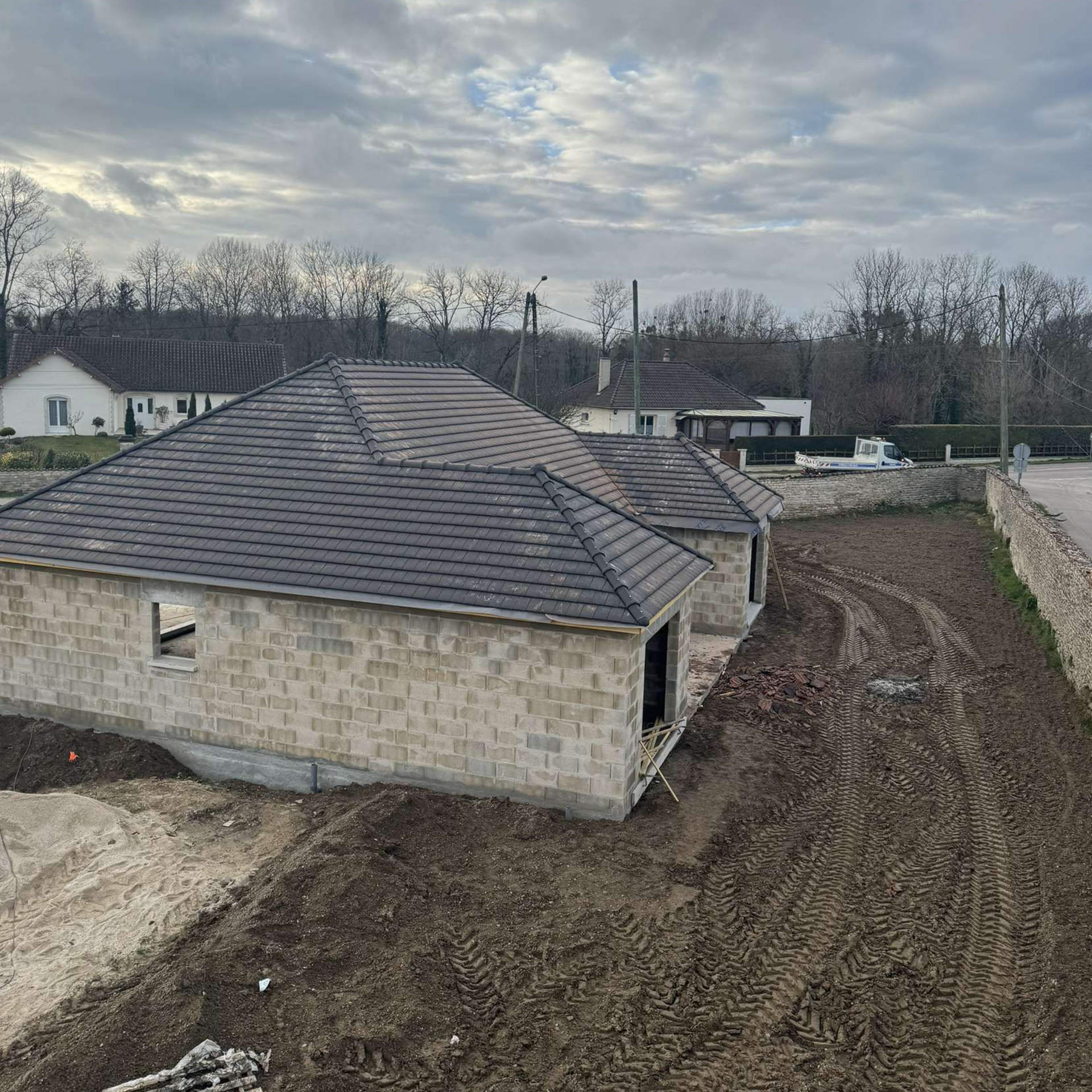 Chantier de construction avec une maison partiellement construite, toit gris, murs en briques et sol en terre battue.