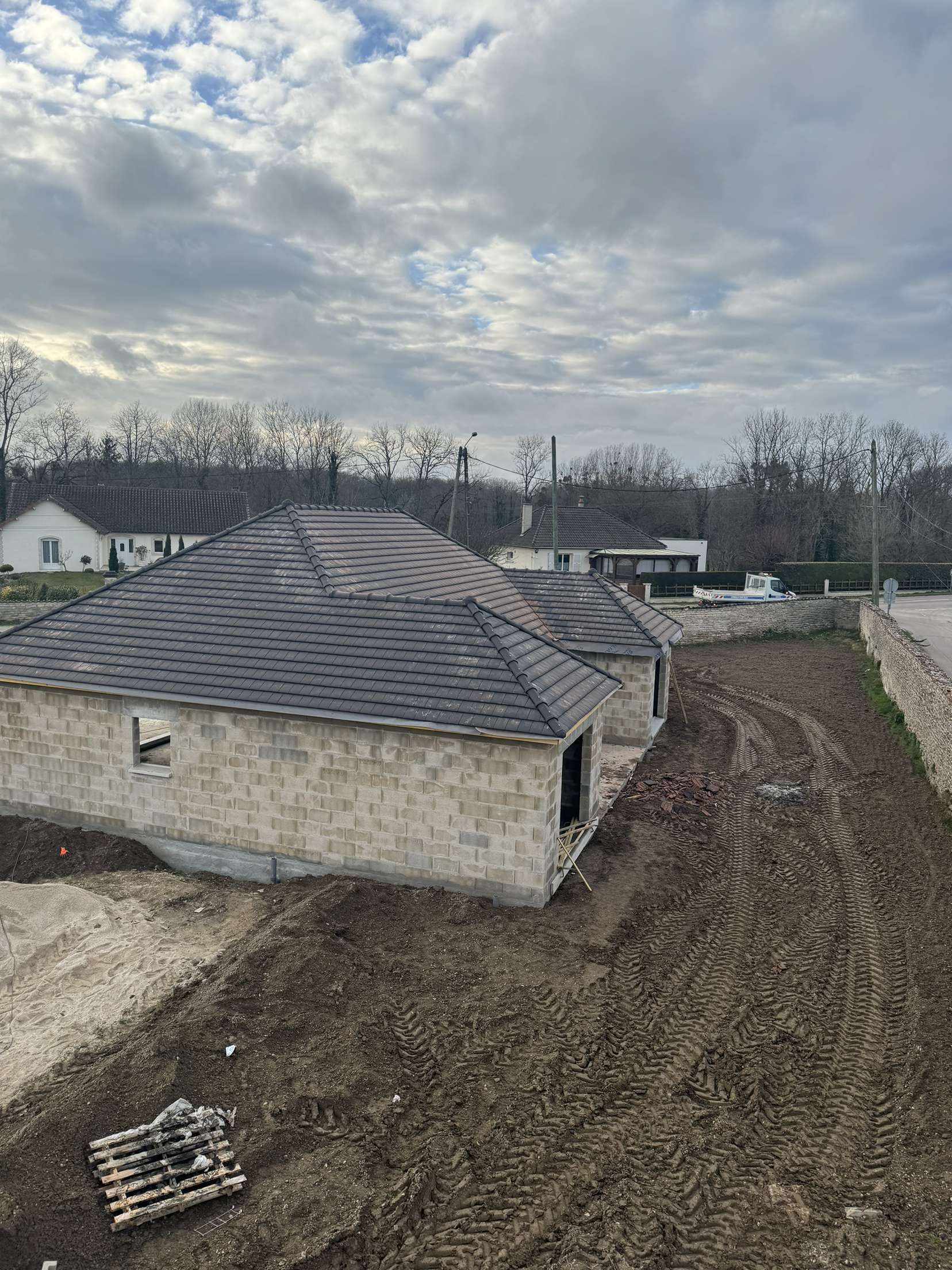 Chantier de construction avec des bâtiments en parpaings beiges, des toits gris et un sol boueux sous un ciel nuageux.