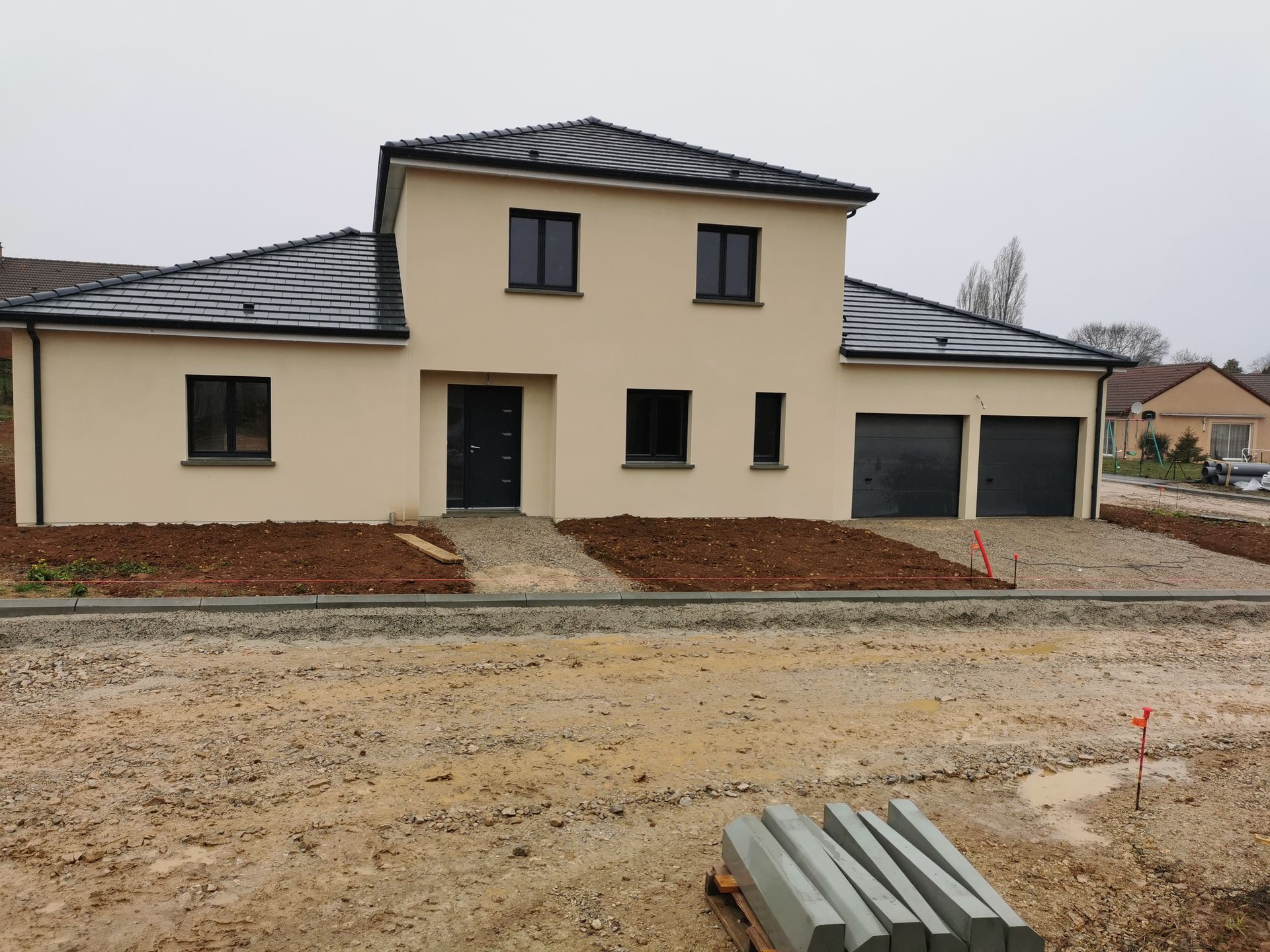 Maison beige à deux étages avec toit noir et garage. Jardin non aménagé à l'avant. Journée nuageuse.