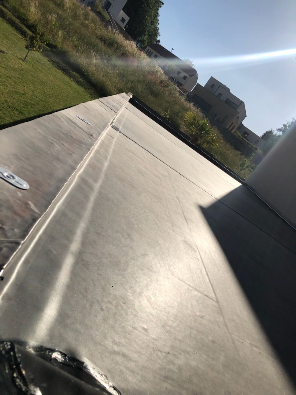 Vue d'un toit plat noir avec un joint. De l'herbe et des bâtiments sont visibles à l'arrière-plan.