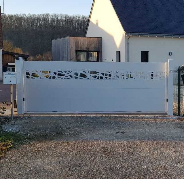 Portail coulissant blanc donnant sur une allée devant une maison moderne, avec un chemin de gravier et des arbres à l'arrière.