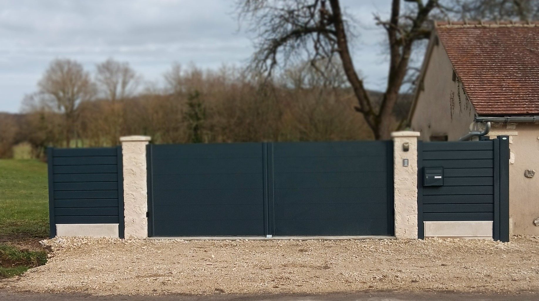 Portail et clôture modernes en métal bleu foncé avec piliers en pierre devant une maison de campagne